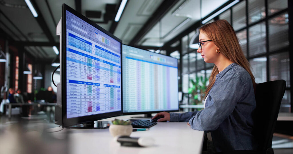 Profissional de óculos e blusa manga longa trabalhando em frente a monitores com dados fiscais.